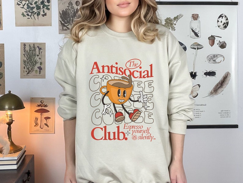K&ouml;nnte beinhalten: Ein beiger Pullover mit einem Grafikdesign, das eine Comic-Kaffeetasse mit dem Text "The Antisocial Club. Espresso yourself, silently." zeigt.
