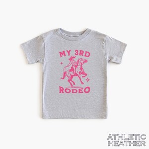 May include: A gray t-shirt with a pink graphic of a girl riding a horse. The text "MY 3RD RODEO" is printed on the shirt.