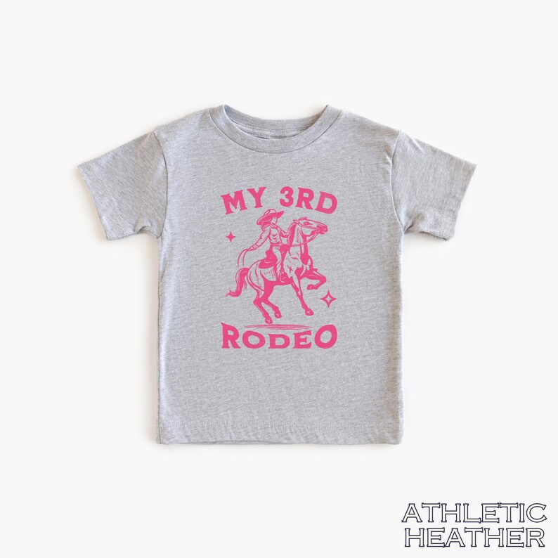 May include: A gray t-shirt with a pink graphic of a girl riding a horse. The text "MY 3RD RODEO" is printed on the shirt.