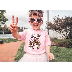May include: A pink t-shirt with a graphic of a girl riding a horse with the text "Lil' Bit Rowdy" on it.