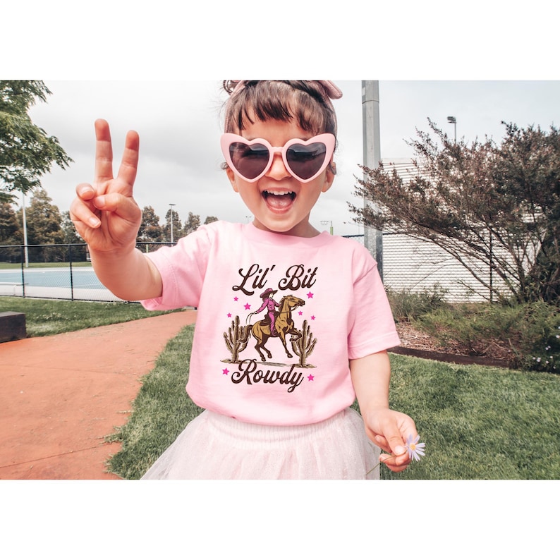 May include: A pink t-shirt with a graphic of a girl riding a horse with the text "Lil' Bit Rowdy" on it.