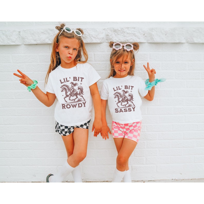 May include: Two young girls wearing matching white t-shirts with a graphic of a cowgirl riding a horse. One shirt says "Lil' Bit Rowdy" and the other says "Lil' Bit Sassy". They are wearing checkered shorts and sunglasses.