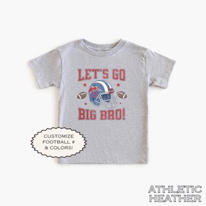 Puede incluir: Camiseta gris jaspeado de manga corta con un dise&ntilde;o de casco de f&uacute;tbol y el texto "LET'S GO BIG BRO!". El dise&ntilde;o incluye un casco azul y rojo, balones de f&uacute;tbol y estrellas. La camiseta tambi&eacute;n tiene el texto "PERSONALIZA F&Uacute;TBOL # & COLORES!"