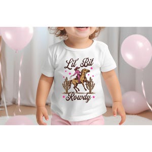May include: White t-shirt with a brown graphic of a girl riding a horse with the text "Lil' Bit Rowdy" in brown letters.