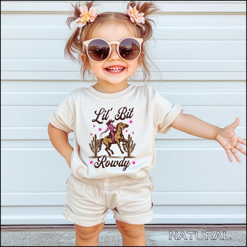 May include: A toddler wearing a beige t-shirt with a graphic of a girl riding a horse and the text "Lil' Bit Rowdy". The toddler is wearing beige shorts and has brown hair with pink flowers in it. The toddler is wearing sunglasses.
