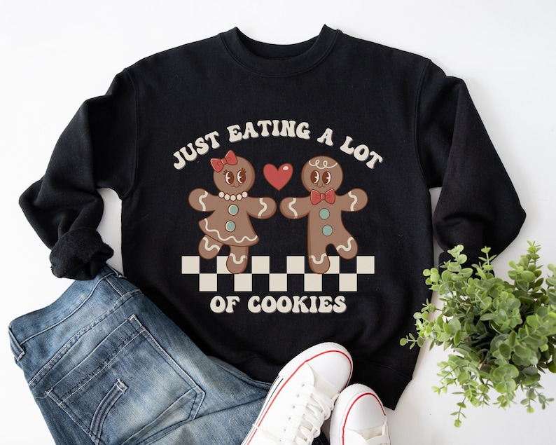 May include: Black sweatshirt with a graphic of two gingerbread people holding hands with a heart between them. The text "Just Eating A Lot Of Cookies" is printed on the sweatshirt.