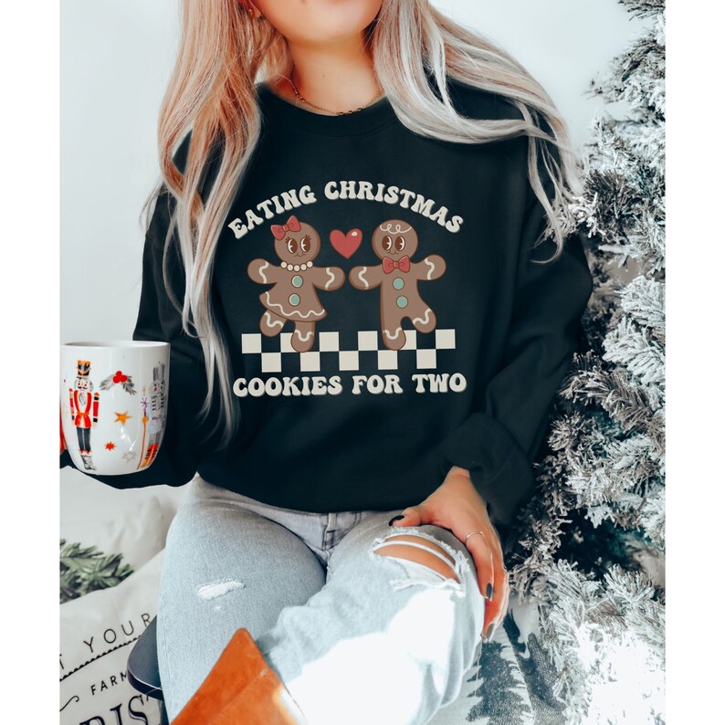 May include: Black sweatshirt with a graphic of two gingerbread people holding hands and a heart. The text reads "Eating Christmas Cookies for Two".