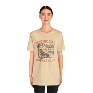 May include: A beige t-shirt with a brown graphic print of a person relaxing on a beach chair under a palm tree. The text "ANTISOCIAL Enjoy the Solitude COFFEE CLUB" is printed on the shirt.