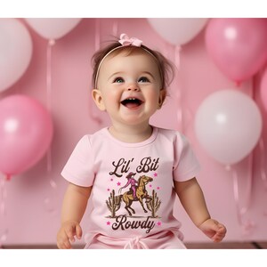 May include: A pink baby t-shirt with a graphic of a girl riding a horse with the text "Lil' Bit Rowdy" in brown letters.