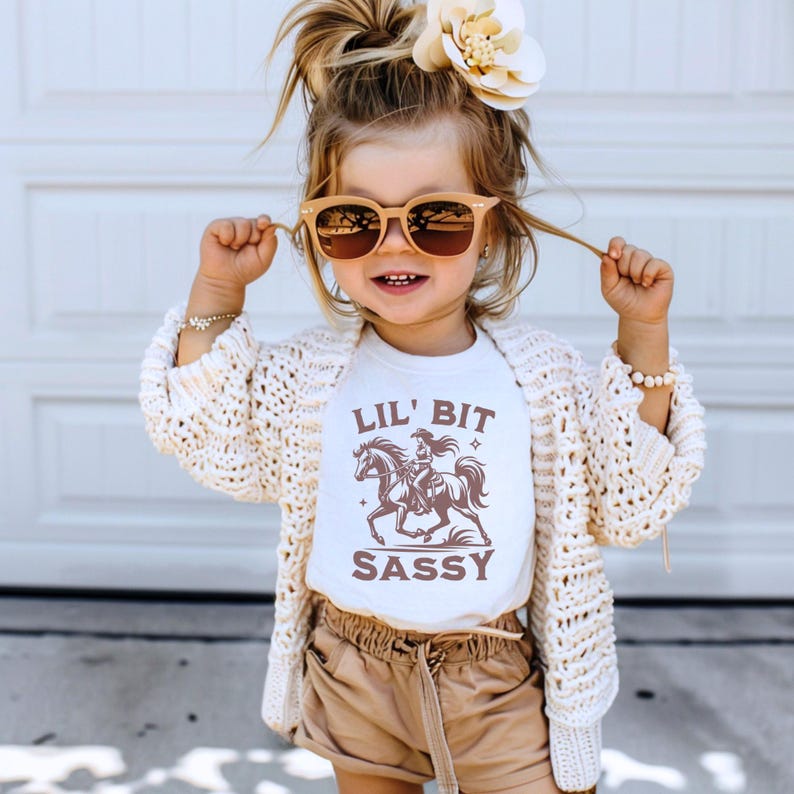May include: A young child wearing sunglasses, a white t-shirt with a horse and rider graphic, and the text "LIL' BIT SASSY". The child is also wearing a cream-colored cardigan and tan shorts. A large flower is in the child's hair.