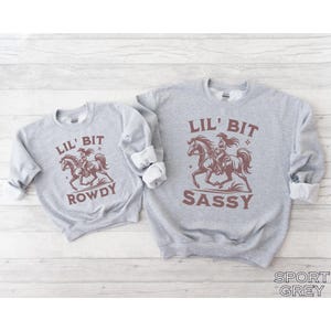 May include: Two gray sweatshirts with rolled-up sleeves. One says "Lil' Bit Rowdy" and the other says "Lil' Bit Sassy." Both have a brown graphic of a cowgirl riding a horse. The bottom right corner of the image says "Sport Grey."