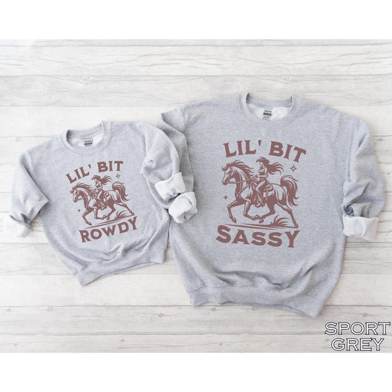 May include: Two gray sweatshirts with rolled-up sleeves. One says "Lil' Bit Rowdy" and the other says "Lil' Bit Sassy." Both have a brown graphic of a cowgirl riding a horse. The bottom right corner of the image says "Sport Grey."