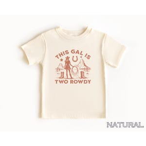 Puede incluir: Una camiseta de color crema con un gr&aacute;fico rojo de una vaquera con un lazo, una herradura y el texto "This Gal Is Two Rowdy".