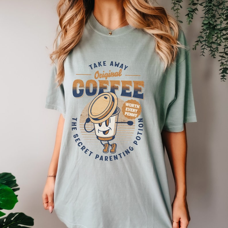 Op de afbeelding: Een lichtblauw t-shirt met een afbeelding van een cartoon koffiekopje met de tekst "Take Away Original Coffee The Secret Parenting Potion Worth Every Penny".