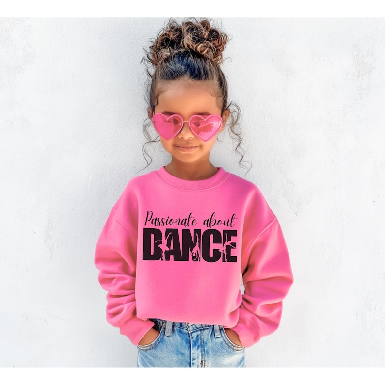 May include: A young girl wearing a pink sweatshirt with the text "Passionate about DANCE" in black. The word "DANCE" is made up of silhouettes of dancers. She is wearing heart-shaped sunglasses and blue jeans.