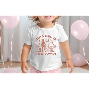 Puede incluir: Camiseta blanca con un gr&aacute;fico marr&oacute;n de una ni&ntilde;a con un sombrero de vaquero sosteniendo un lazo. El texto "This gal is two rowdy" est&aacute; impreso debajo del gr&aacute;fico.