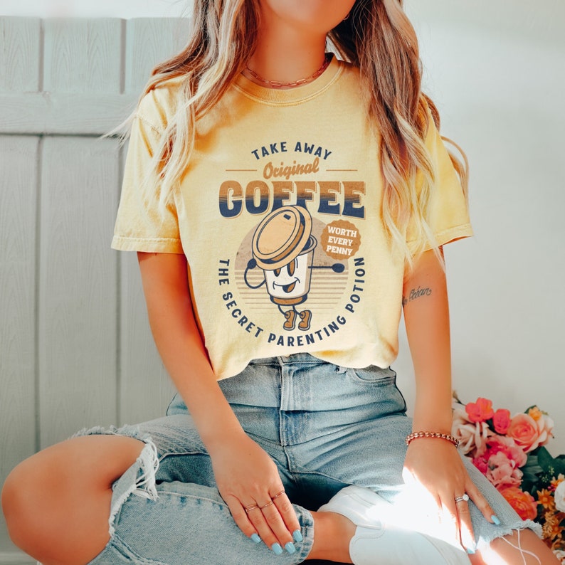 Op de afbeelding: Een geel T-shirt met een retro-grafische afbeelding van een koffiekopje met de tekst "Take Away Original Coffee The Secret Parenting Potion Worth Every Penny".
