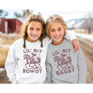 May include: Two girls wearing matching sweatshirts. One is white with "LIL' BIT ROWDY" and a graphic of a cowgirl on a horse. The other is gray with "LIL' BIT SASSY" and the same graphic. The girls are smiling.
