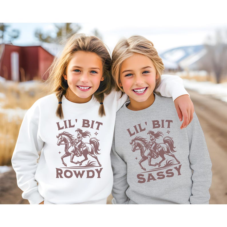 May include: Two girls wearing matching sweatshirts. One is white with "LIL' BIT ROWDY" and a graphic of a cowgirl on a horse. The other is gray with "LIL' BIT SASSY" and the same graphic. The girls are smiling.