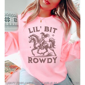 May include: Light pink sweatshirt with a graphic of a cowgirl riding a horse, the words "LIL' BIT ROWDY" are above and below the image. The text "LIGHT PINK-ONLY AVAILABLE IN ADULT SIZES" is at the bottom of the image.