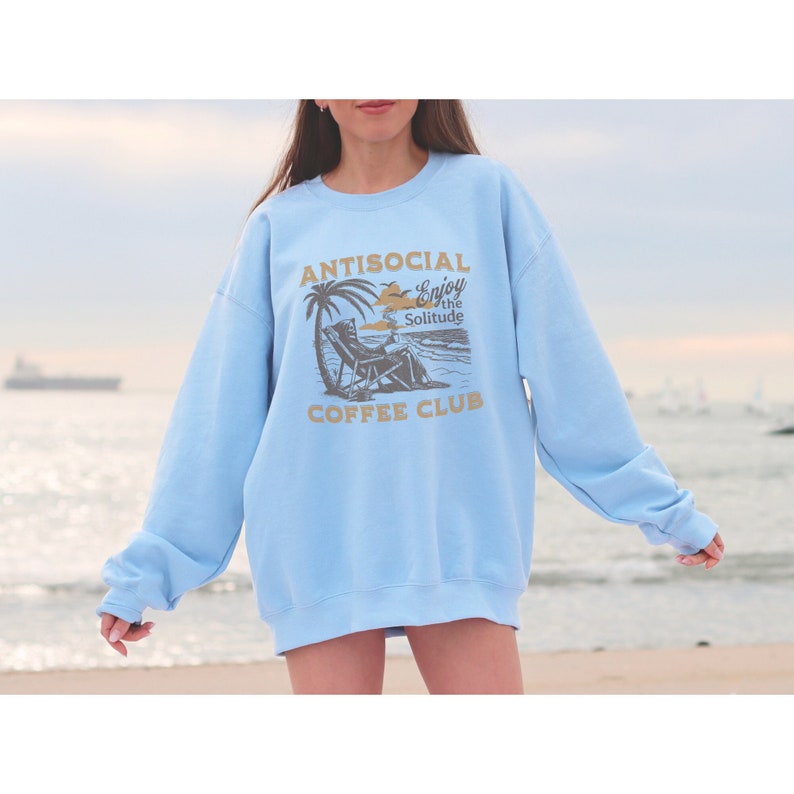 May include: Light blue sweatshirt with a graphic print of a person sitting on a beach chair under a palm tree. The text on the sweatshirt reads "Antisocial Enjoy the Solitude Coffee Club".