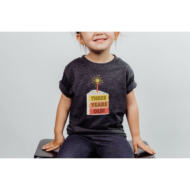 May include: A dark grey t-shirt with a colorful birthday cake graphic that says "THREE YEARS OLD!"