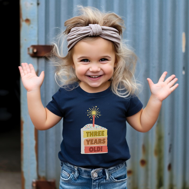 May include: A young child wearing a navy blue t-shirt with a birthday cake graphic and the text "THREE YEARS OLD!" The child is smiling and has blonde hair with a light brown headband.