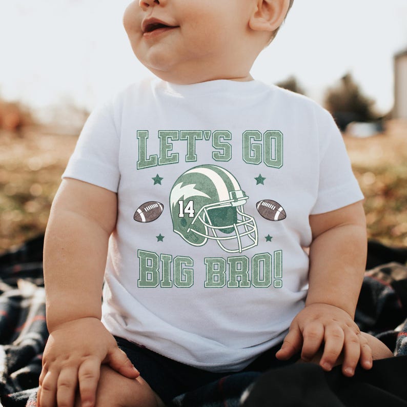 Puede incluir: Camiseta blanca con texto y gr&aacute;ficos verdes. La camiseta dice "LET'S GO BIG BRO!" y presenta un casco de f&uacute;tbol con el n&uacute;mero 14, junto con dos balones de f&uacute;tbol y estrellas. Un beb&eacute; lleva la camiseta.