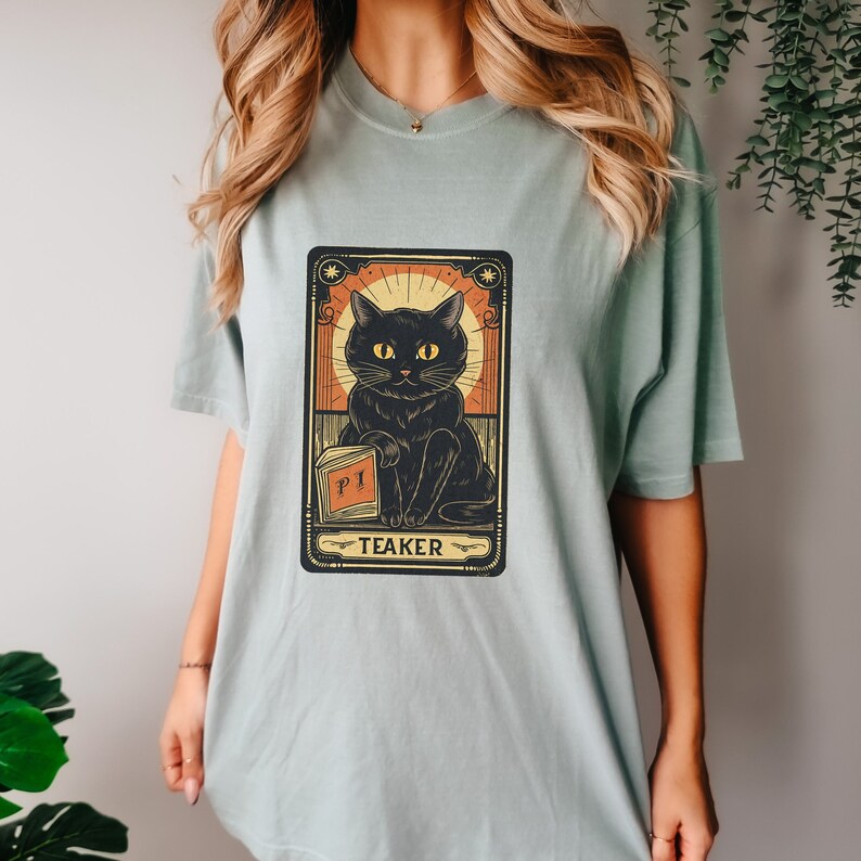 May include: A light blue t-shirt with a black cat illustration in a vintage tarot card style. The card reads "Teaker" and features a black cat holding a book that says "PI".