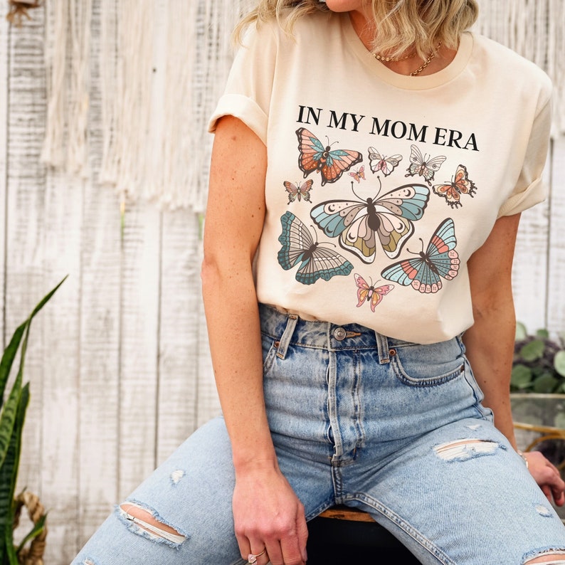 K&ouml;nnte beinhalten: Ein beiges T-Shirt mit dem Aufdruck "IN MY MOM ERA" in Schwarz. Das Shirt zeigt ein farbenfrohes Schmetterlingsdesign mit mehreren Schmetterlingen in verschiedenen Blaut&ouml;nen, Rosa und Orange.