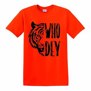 Puede incluir: Camiseta naranja brillante con un dise&ntilde;o de tigre negro y las palabras "WHO DEY". La cara del tigre est&aacute; a la izquierda, con el texto alineado verticalmente a la derecha. La camiseta es de cuello redondo.