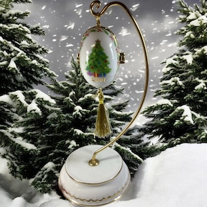 May include: A white egg-shaped Christmas ornament with a painted Christmas tree and the year 2001. The ornament hangs from a gold-colored metal stand with a tassel. The base of the stand is white with gold trim.