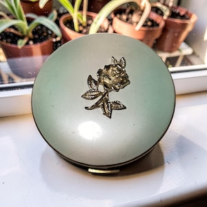 May include: A round, pale green compact with a gold-colored rose and leaf design on the lid. The compact has a gold-colored rim and a small clasp. The background includes potted plants near a window.