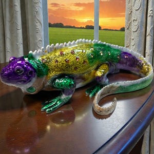 May include: A decorative lizard figurine in Mardi Gras colors: purple, green, and gold. The lizard has a curved tail and is embellished with small red gems. It sits on a dark brown wooden table, with a sunset visible in the background.