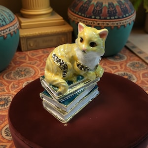 May include: A yellow cat figurine with a white chest and sparkling accents, perched atop a stack of teal and gold-edged books. The cat is in a relaxed pose, adding a touch of whimsy to the decorative piece.