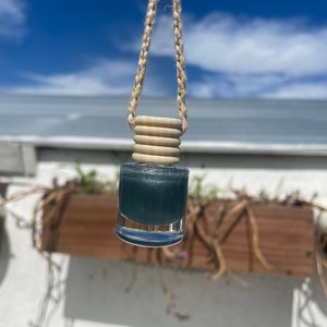 May include: A small, clear glass bottle filled with a teal-colored liquid, suspended by a braided rope. The bottle has a wooden cap and is hanging against a backdrop of a blue sky and a white building.