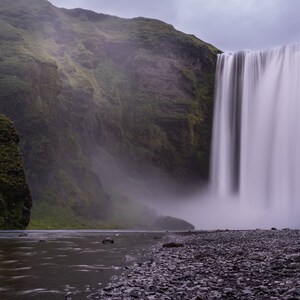 May include: A powerful waterfall cascades down a rocky cliff face, creating a misty spray. Lush green vegetation covers the surrounding landscape.