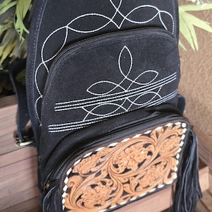 Puede incluir: Mochila de gamuza negra con costuras blancas y una solapa de cuero marr&oacute;n con relieve y flecos.