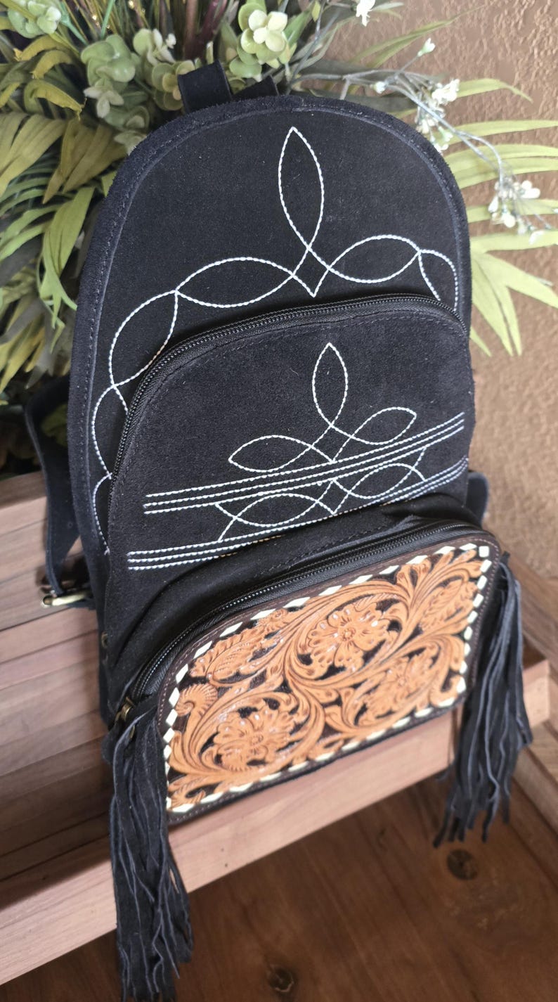 Puede incluir: Mochila de gamuza negra con costuras blancas y una solapa de cuero marr&oacute;n con relieve y flecos.