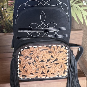 Puede incluir: Mochila de gamuza negra con costuras blancas y una solapa de cuero marr&oacute;n con dise&ntilde;os florales. La mochila tiene flecos negros en la parte inferior.