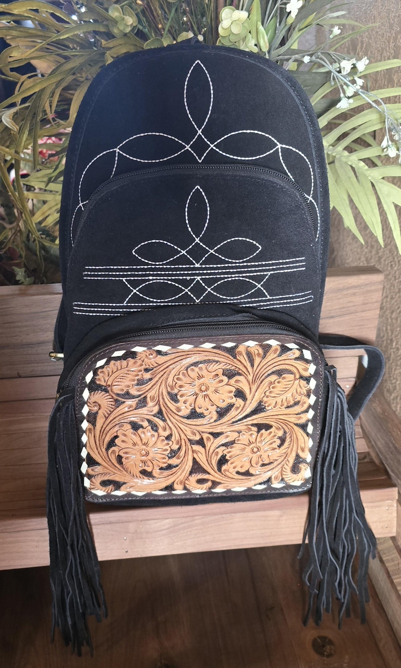 Puede incluir: Mochila de gamuza negra con costuras blancas y una solapa de cuero marr&oacute;n con dise&ntilde;os florales. La mochila tiene flecos negros en la parte inferior.