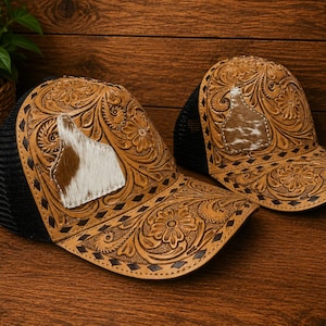 Può includere: Due cappellini da camionista in pelle marrone con intricati strumenti floreali e geometrici. Ogni cappello presenta una toppa in pelle di mucca con un motivo bianco e marrone. I cappelli hanno schienali in rete nera e sono esposti su una superficie di legno.