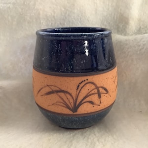 May include: A handmade ceramic stemless wine glass. The top and bottom are a speckled dark blue, with a band of tan in the middle. The tan band features a brown floral design. The glass is approximately 4 inches tall.