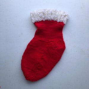May include: A small, red knitted Christmas stocking with a white, fluffy cuff. The stocking is made of a textured yarn and is likely handmade. The overall design is simple and classic, perfect for holiday decor.