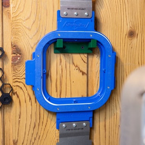 May include: A blue Mighty Hoop embroidery hoop is mounted on a wooden surface. The hoop has a rectangular shape with a white inner border and metal attachments. The text "MH-MT400-55-BKT" is visible on the metal parts.
