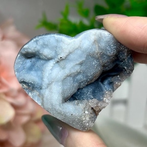 May include: A heart-shaped crystal geode with a mix of blue, white, and gray hues. The stone has a rough, textured surface with sparkling crystal formations. The geode is held up by a hand.