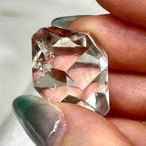 Puede incluir: Una piedra preciosa transparente y facetada con un corte geométrico de múltiples lados. La piedra es incolora, con bordes afilados y superficies planas que reflejan la luz. Presenta pequeñas inclusiones oscuras visibles en su interior.