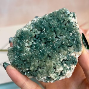 Könnte beinhalten: Ein Cluster aus teal-farbenen Kristallen mit einer rauen, strukturierten Oberfläche. Die Kristalle sind dicht gepackt und erzeugen einen funkelnden Effekt. Die Basis des Clusters ist hell, cremefarben.