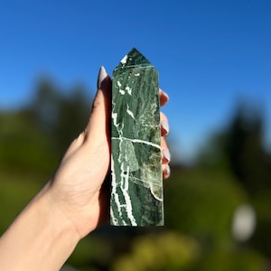 Può includere: Un obelisco di pietra verde e bianca è tenuto contro un cielo blu. L'obelisco è rettangolare con una sommità appuntita e presenta un motivo marmorizzato di verde scuro e bianco. La pietra è liscia e lucidata.