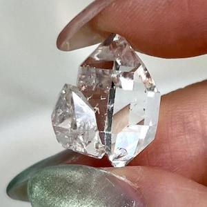 Peut inclure: Un petit cristal de quartz transparent à double terminaison avec des facettes naturelles et des inclusions internes. La pierre présente une pointe hexagonale principale avec un cristal secondaire plus petit attaché sur le côté, offrant une grande transparence.
